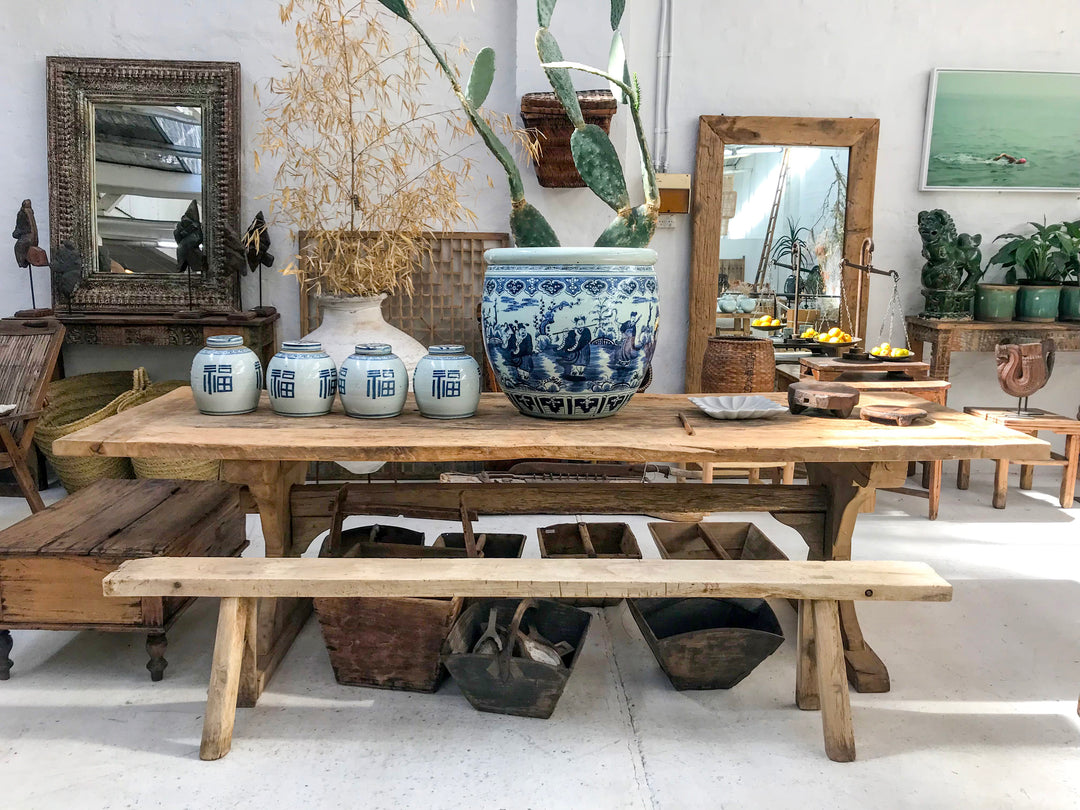 A rustic wooden dining table with matching benches in a styled home decor showroom, decorated with blue and white ceramic jars, a large blue patterned pot, and dried plants.