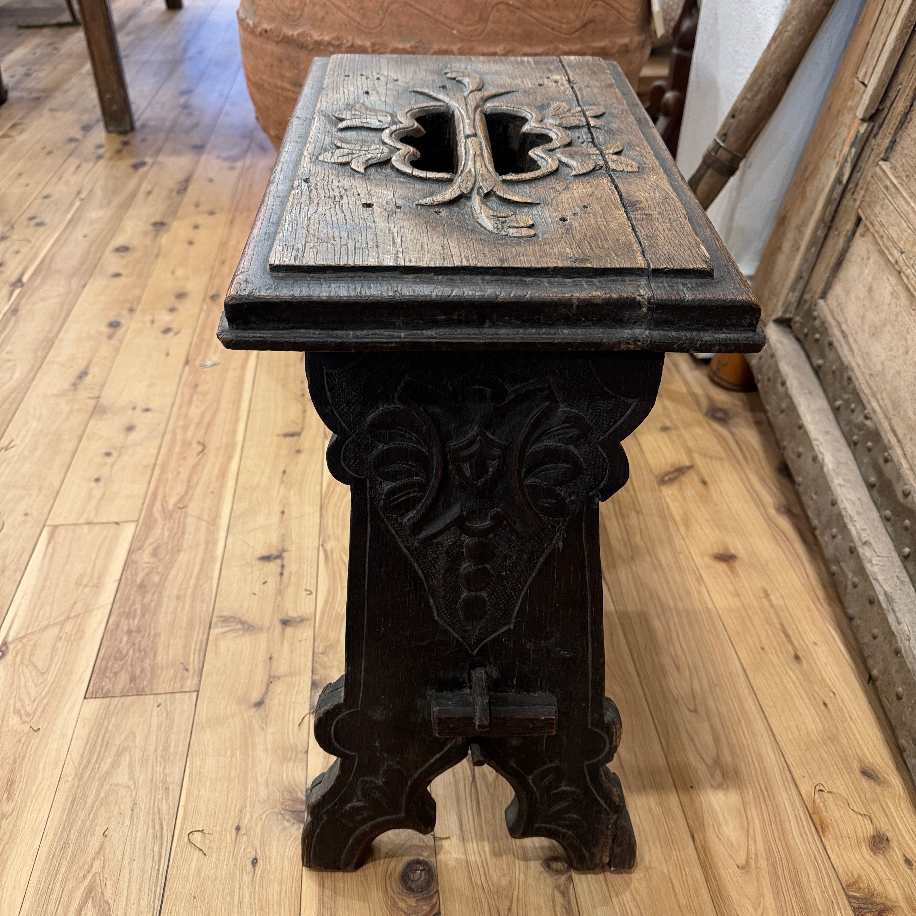 An antique dark carved wooden stool or table with a rectangular top, ornate floral relief details, and a decorative pedestal base, photographed on a light wood floor in an indoor room.