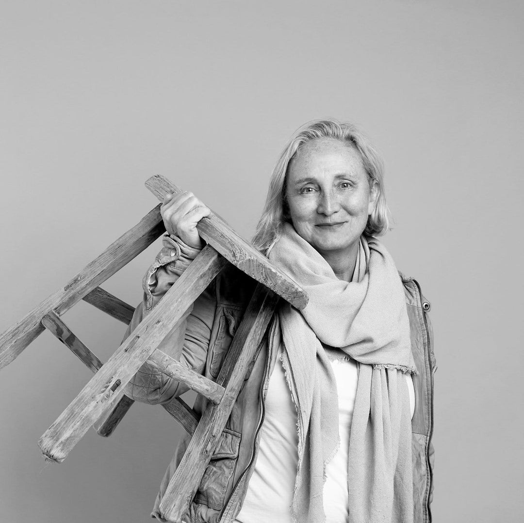 A black and white portrait of Kathrin Bruce, a woman holding a rustic wooden stool against a plain gray background.