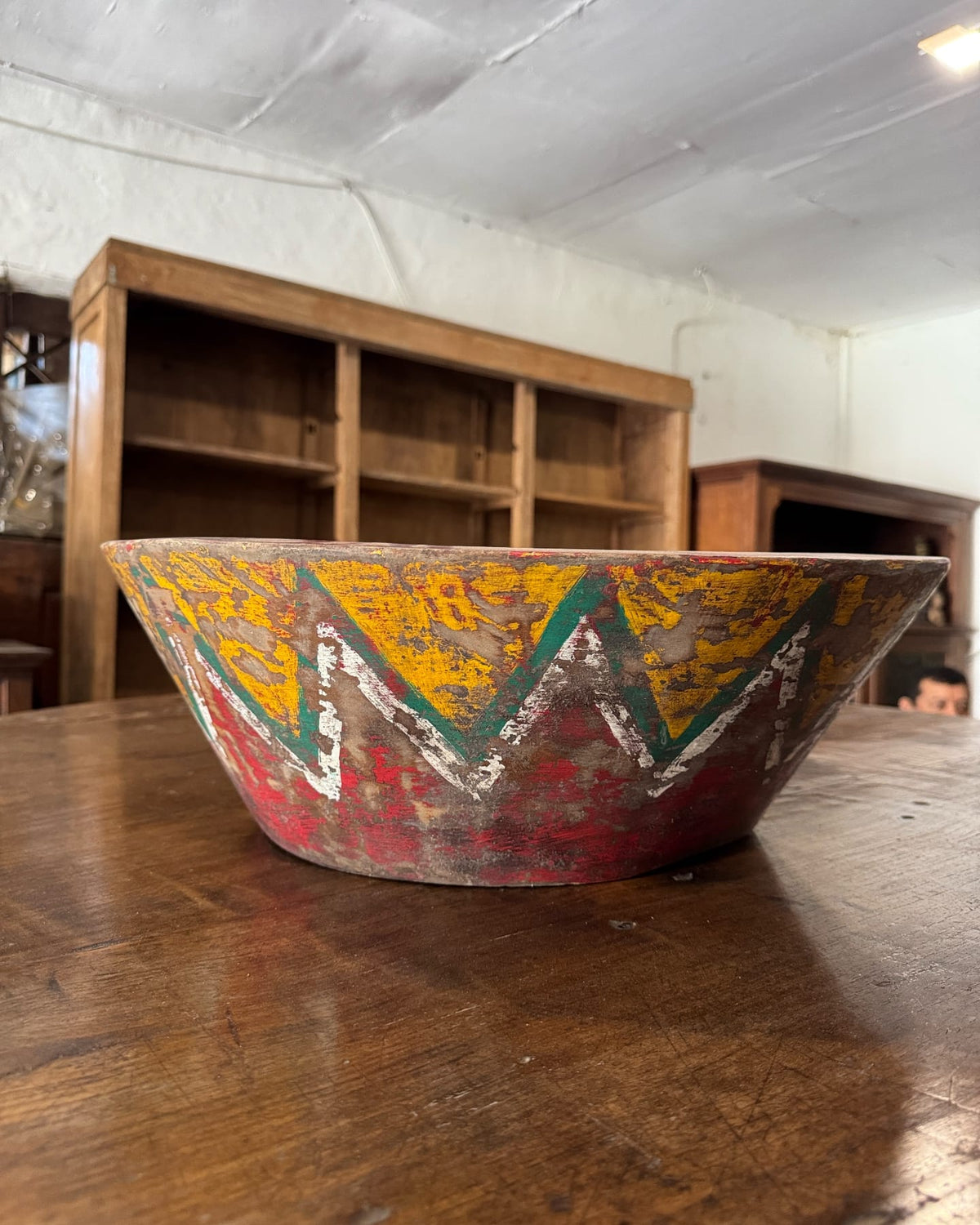 A large, rustic hand-painted wooden bowl with a colorful geometric pattern, resting on a wooden table 